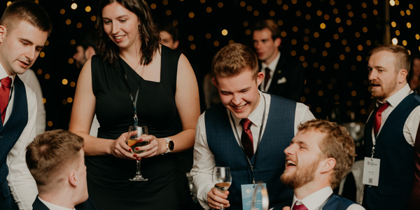 Students standing and sitting around a table laughing. Some are holding drinks.