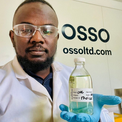 Mohaned Aboshatta in a lab, wearing blue gloves and protective glasses, holding a bottle of clear fluid.