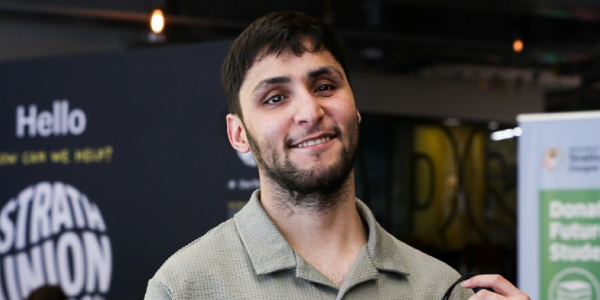 Abdul, a young man with short dark hair, beard and moustache, smiling inside the Strath Union building. There are signs behind him that say Hello, welcome to Strath Union. How can we help?