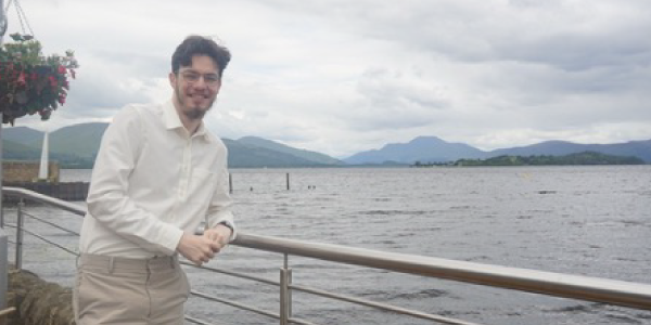 Duncan, a young man with short brown hair, a beard, and wearing glasses, leaning against a railing overlooking a loch with hills in the distance. Duncan is wearing a smart shirt and trousers.