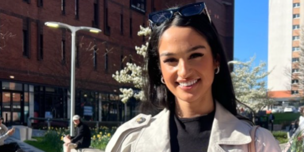 Sharan, a young woman with long dark hair and wearing professional clothes, smiling on campus outside on a sunny day. The Lord Hope building is behind her.